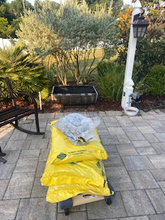 In the forefront, there is a flat cart with wheels, holding 3 large yellow bags of dirt, and a clear plastic bag with rocks in it. Behind this is a half Whiskey barrel made into a plant trough. There is also a white lamp post with a black hanging lamp. There are sandstone colored pavers, that go up to mulch and some bushes. 