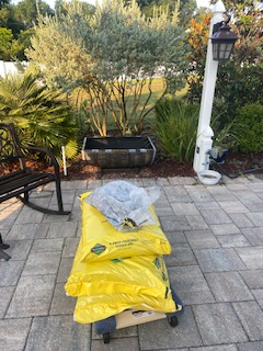 In the forefront, there is a flat cart with wheels, holding 3 large yellow bags of dirt, and a clear plastic bag with rocks in it. Behind this is a half Whiskey barrel made into a plant trough. There is also a white lamp post with a black hanging lamp. There are sandstone colored pavers, that go up to mulch and some bushes.