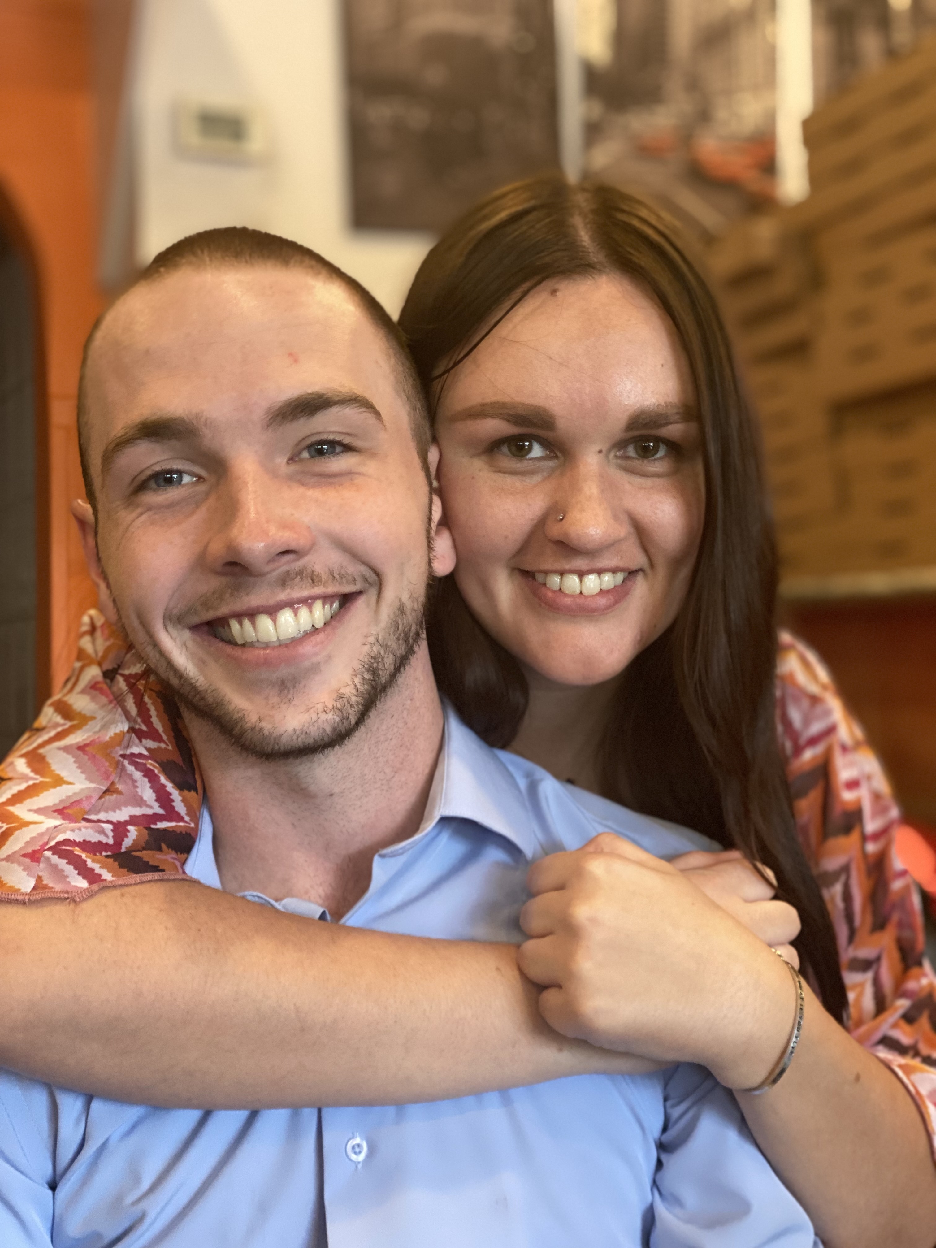 Michael is in a blue button shirt with a smile, slight beard and mustache. Jorjia is standing behind him with her right arm across his chest & her hands crossed on his left shoulder. Her brown hair is straight. They are both smiling. 