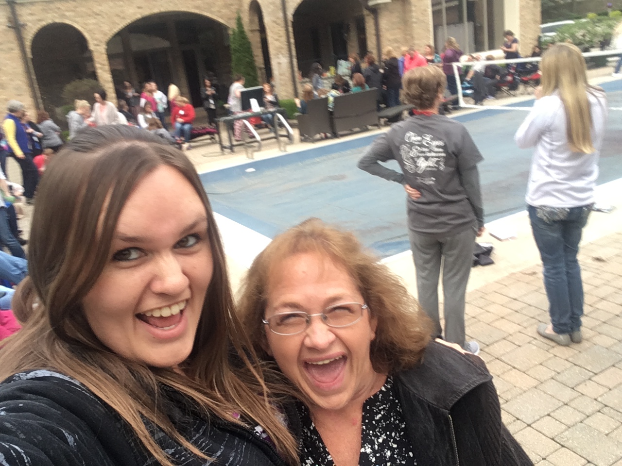 Jorjia and Amy in winter coats standing on a pool deck with their mouths open like they are screaming with excitement.  There are people along the pool, as well as 2 ladies standing behind them with their back to them