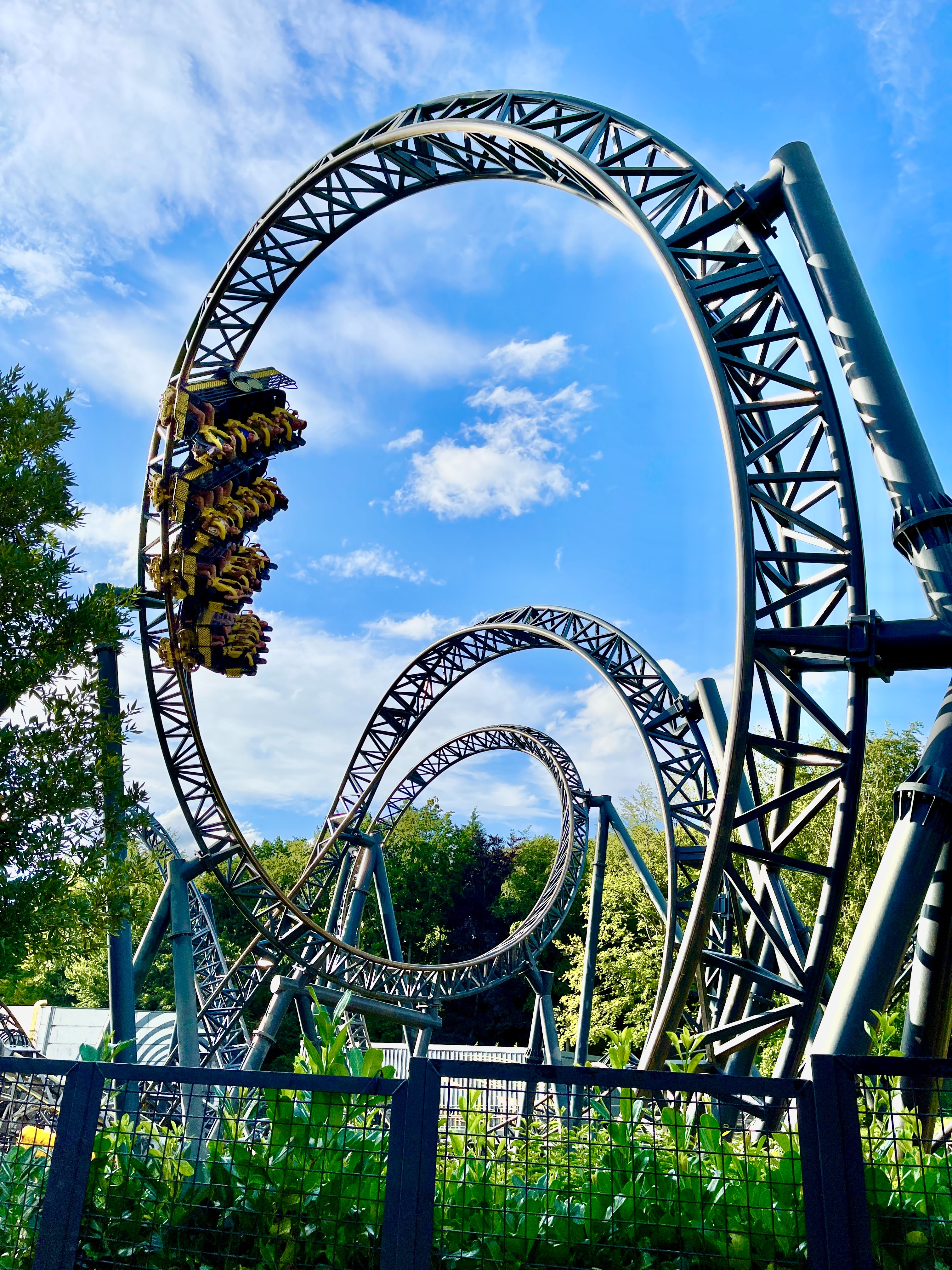 Twisting and turning rollercoaster with in the distance people in a yellow car upside down also a beautiful blue sky with fluffy white clouds behind it and you can also see trees and green bushes 