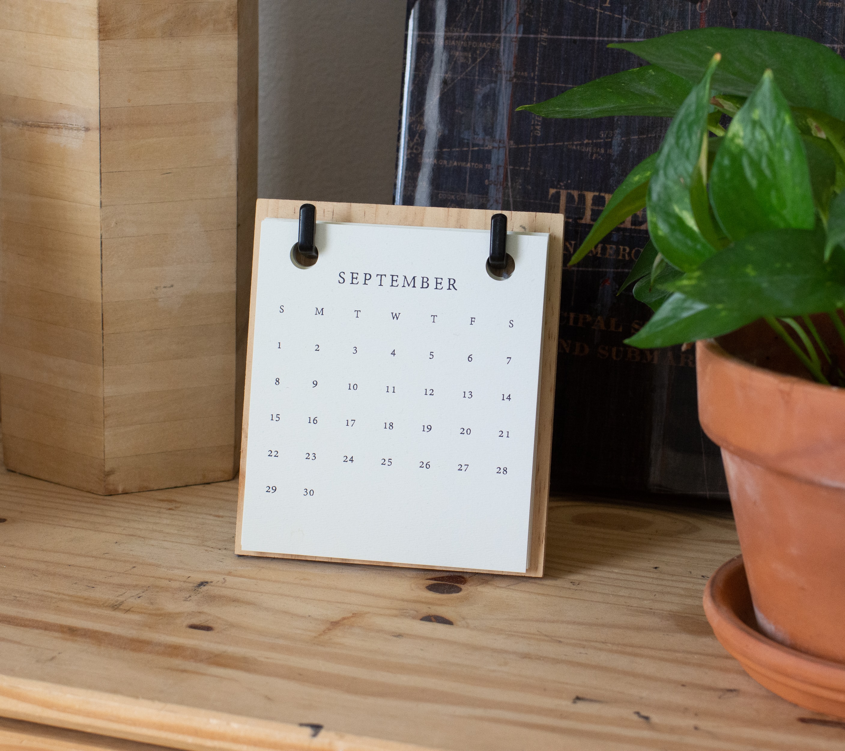 Calendar of September, plant in a planter, both on a wooden desk or shelf.