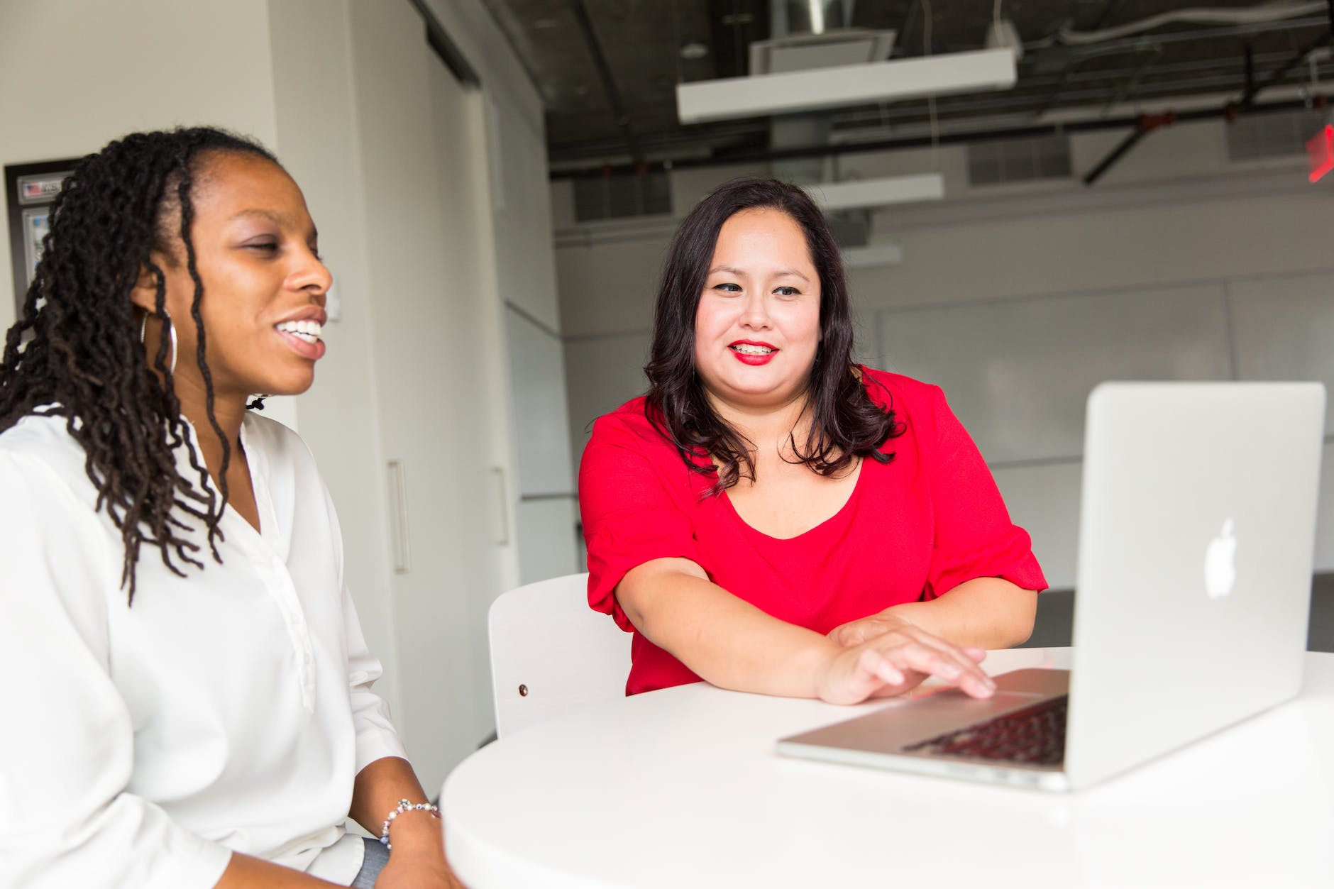 Two females at a table with an Apple computer - one seems to be helping the other. 