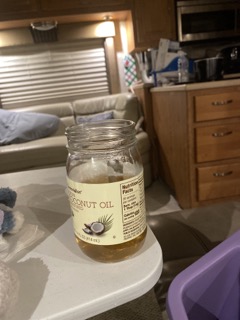 small glass jar with a coconut oil label, about half full of an amber liquid. Sitting on the corner of a plastic table. Background shows interior of the coach, as well as a bit of a plastic lap tray. 
