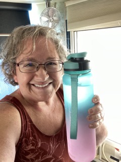 Amy holding her 32 ounce refillable water bottle, standing by the door of the coach. She is smiling and wearing a tank top and glasses. 
