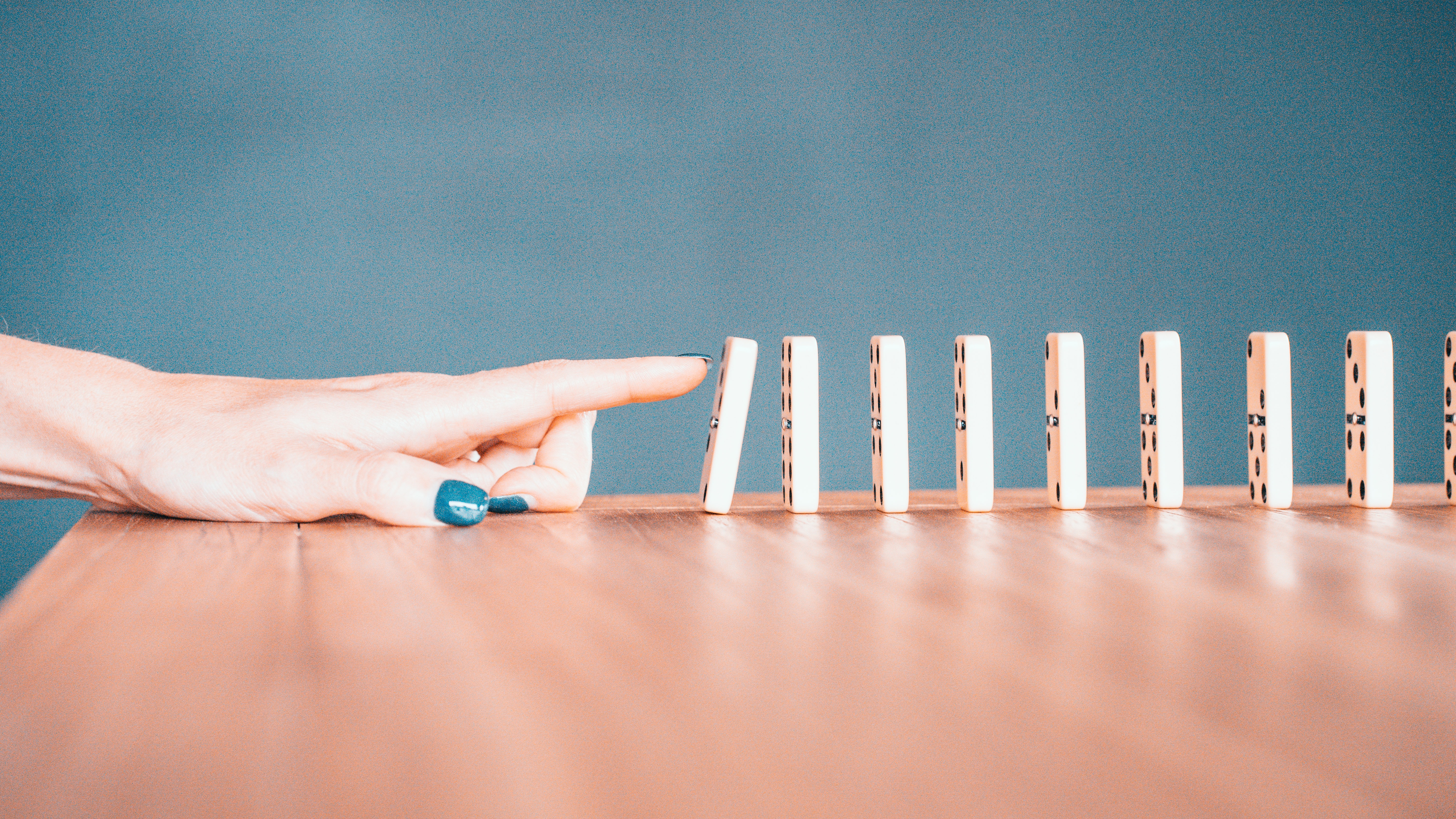 dominos standing up in a line on a wooden table, with a woman's hand on the table with her pointer finger reaching and knocking the first domino