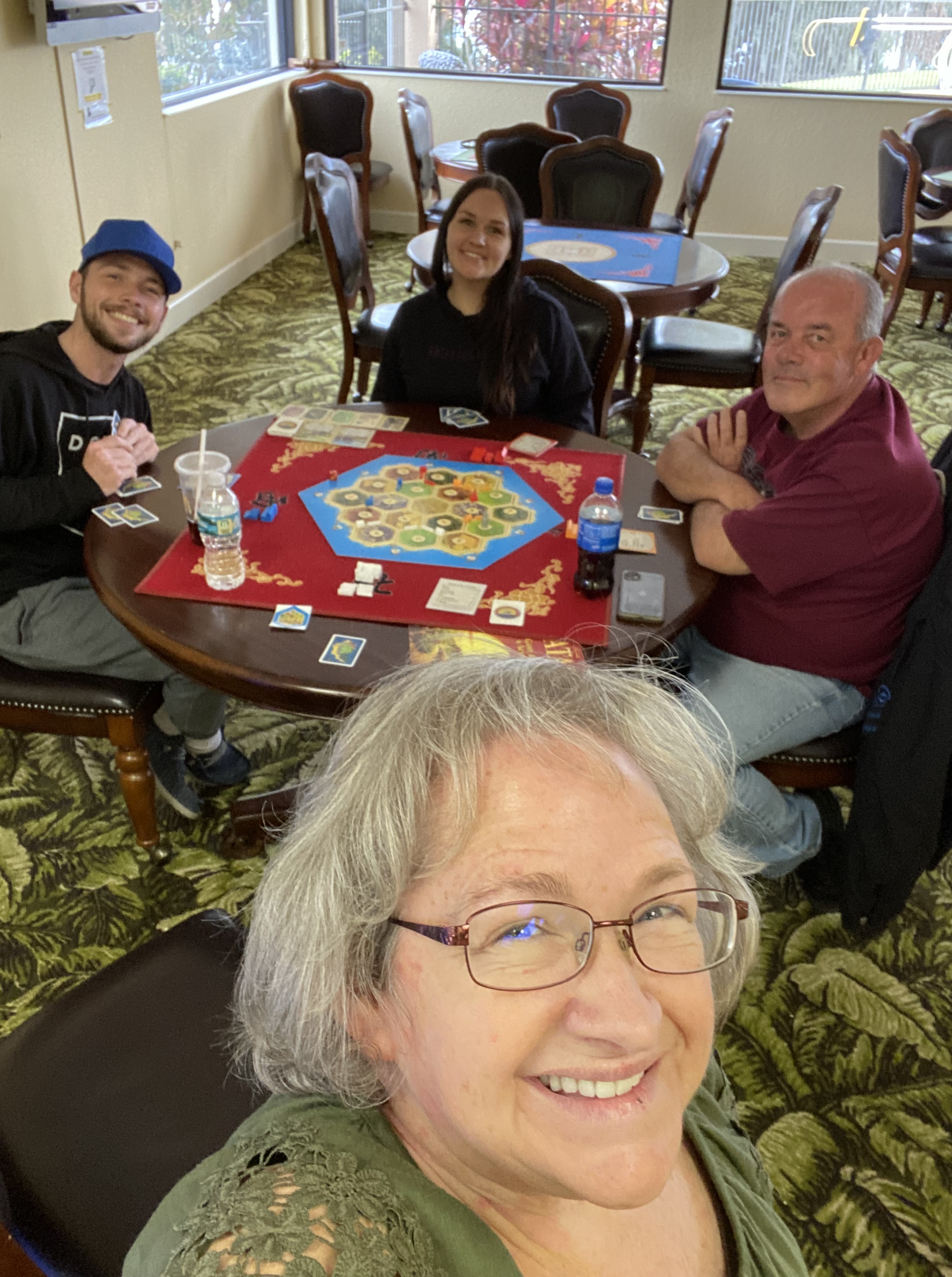 Amy, Ken, Jorjia, and Michael at a table playing Catan, the board game. In the clubhouse, with other tables in the background. 3 windows behind them. 