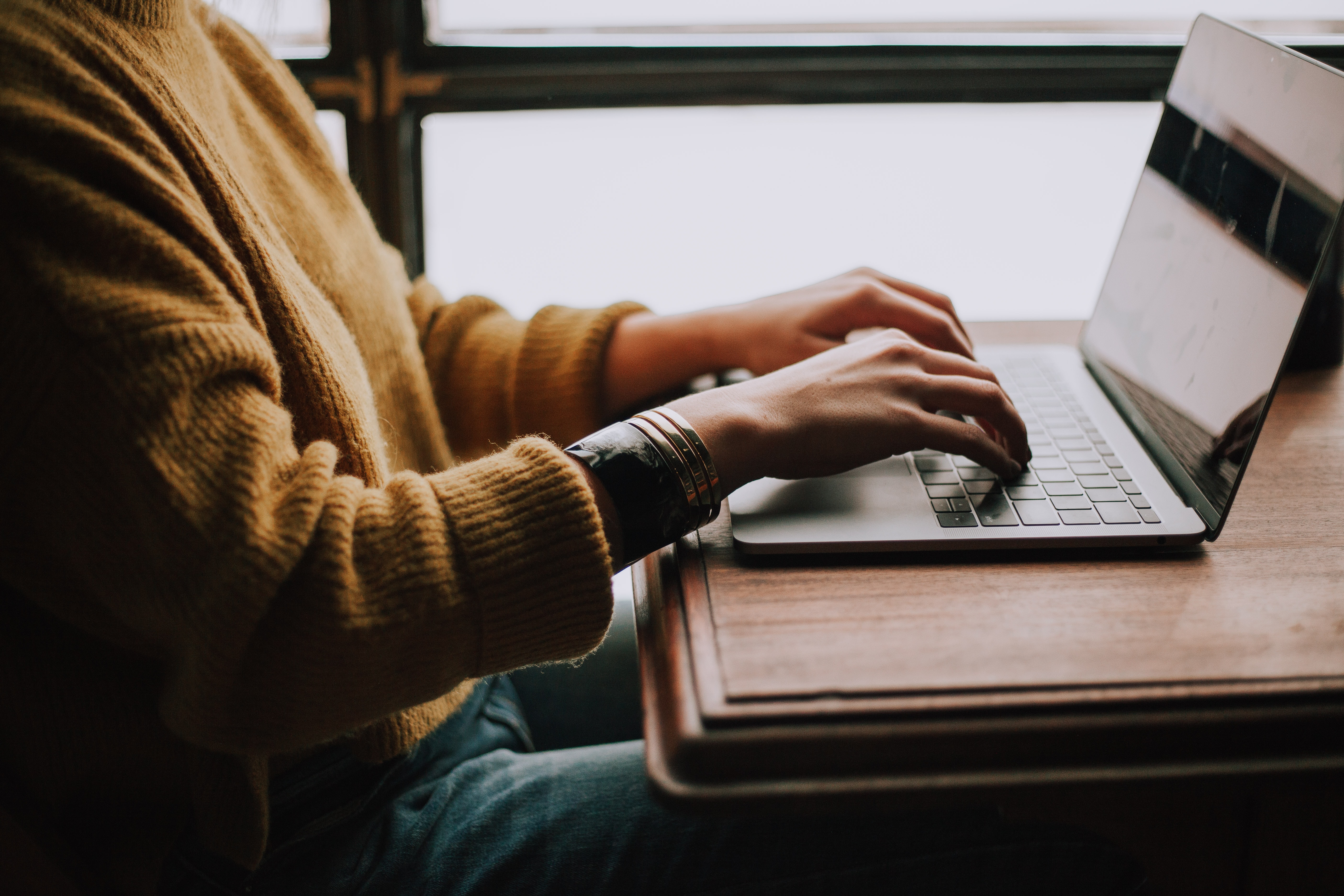 Photo of a woman in a sweater at a computer on a table near a window
  